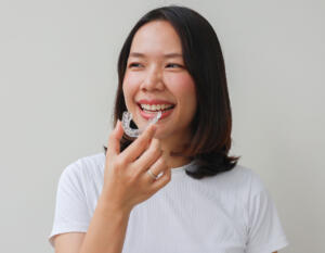 Eine junge Frau vor hellgrauem Hintergrund setzt eine transparente Zahnschiene ein und lächelt.